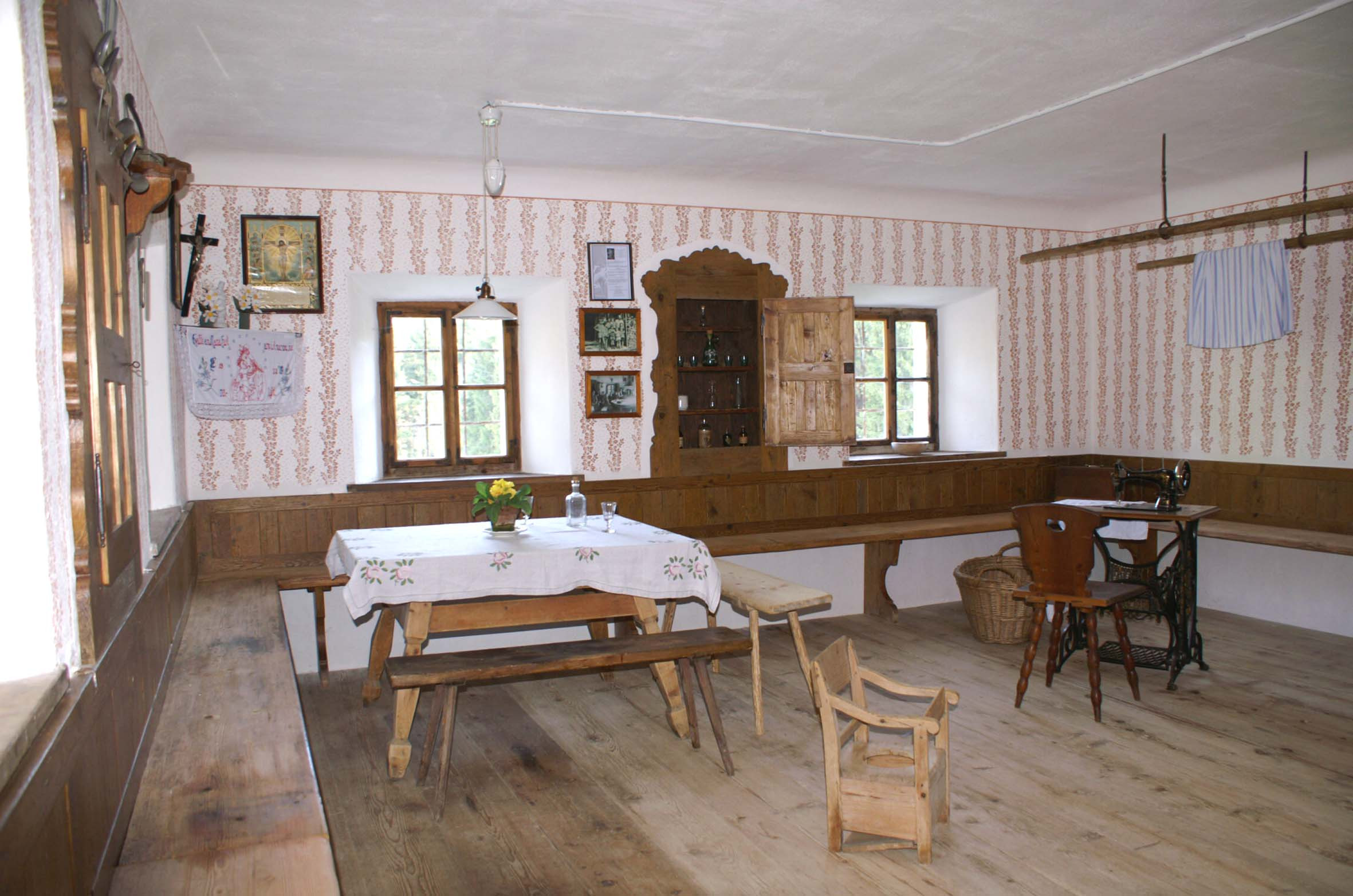 Wohnstube mit umlaufender Bank, rundem Tisch, Tretnähmaschine und Walzmustern an den Wänden